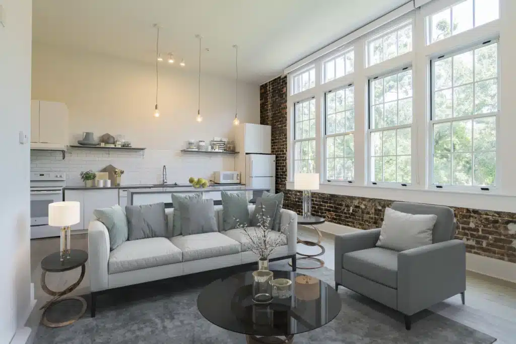 Modern open-plan living room and kitchen with a gray sofa, armchair, round coffee tables, large windows, exposed brick, and white cabinetry.