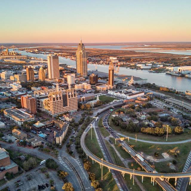Aerial view of downtown Mobile, Alabama, showing office buildings, highways, and the Mobile River at sunset.