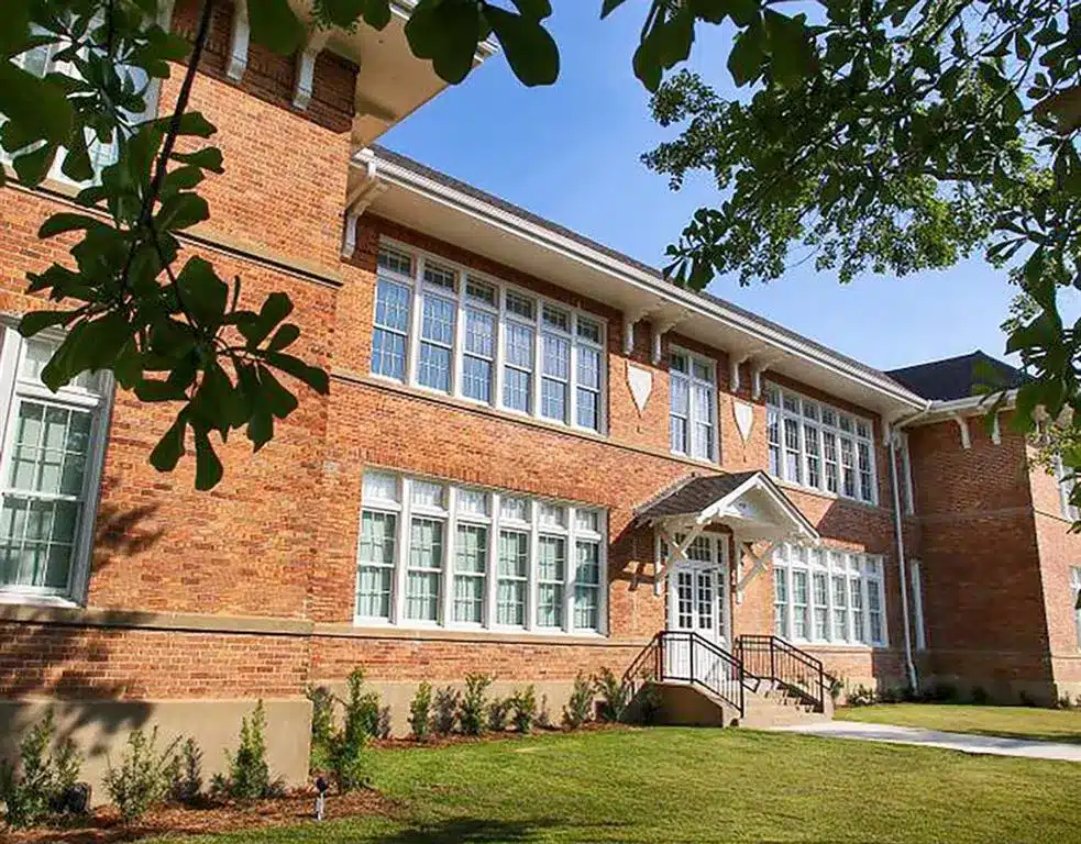 A two-story brick building with many windows and a white entrance, surrounded by green grass and trees under a blue sky.