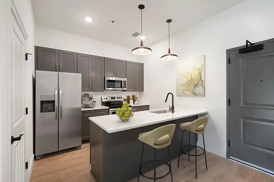 Modern kitchen with gray cabinets, stainless steel appliances, a white island with sink, two beige barstools, pendant lights, and a wall art piece near a gray door.